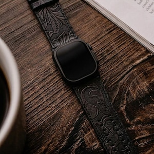 Può includere: Smartwatch nero con cinturino in pelle nera goffrata. Il cinturino presenta un motivo floreale dettagliato e una fibbia in metallo. L'orologio è appoggiato su una superficie di legno accanto a una rivista.