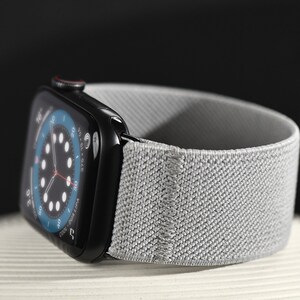 May include: A black Apple Watch Series 8 with a gray sport loop band. The watch face displays a circular analog clock with a blue background.