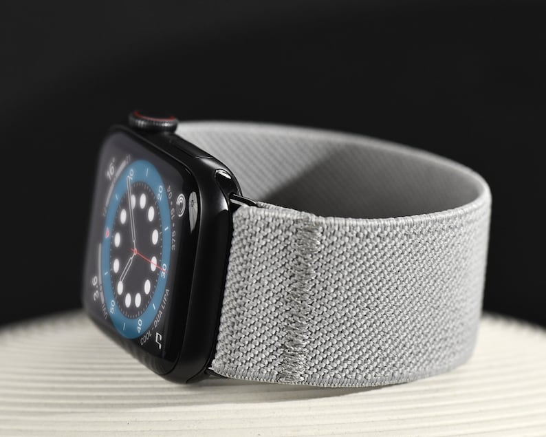 May include: A black Apple Watch Series 8 with a gray sport loop band. The watch face displays a circular analog clock with a blue background.