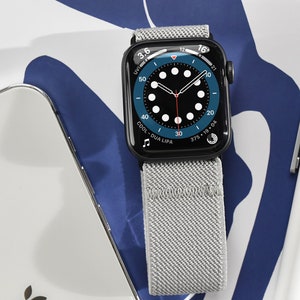 May include: A black Apple Watch with a gray sport loop band. The watch face is a blue analog clock with white markings and a red second hand. The text "COOL - DUA LIPA" is displayed on the watch face.