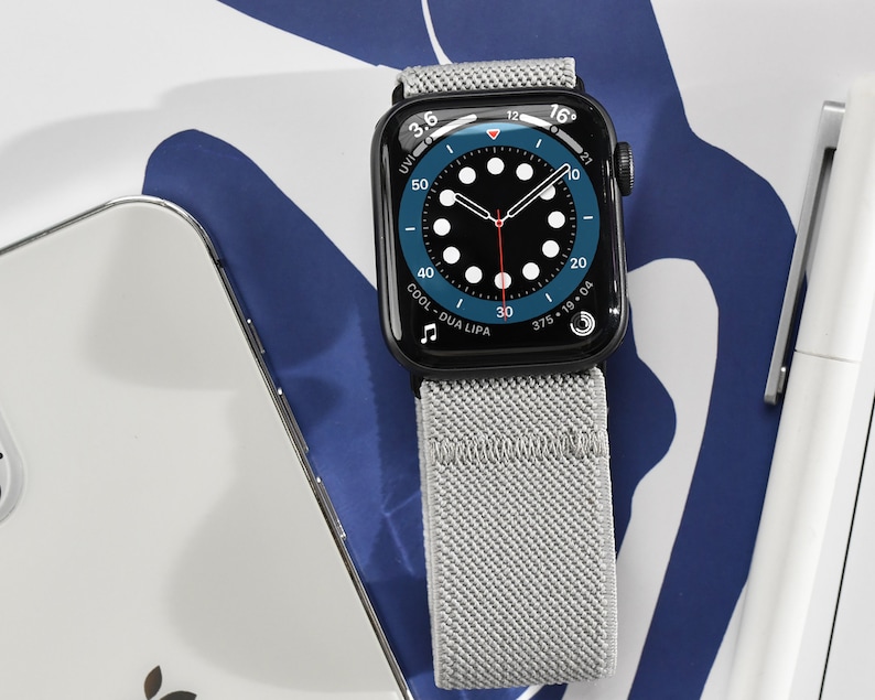 May include: A black Apple Watch with a gray sport loop band. The watch face is a blue analog clock with white markings and a red second hand. The text "COOL - DUA LIPA" is displayed on the watch face.