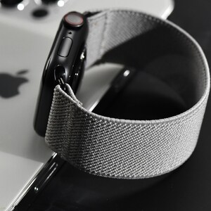 May include: A black Apple Watch with a gray sport loop band. The watch is on a white surface.
