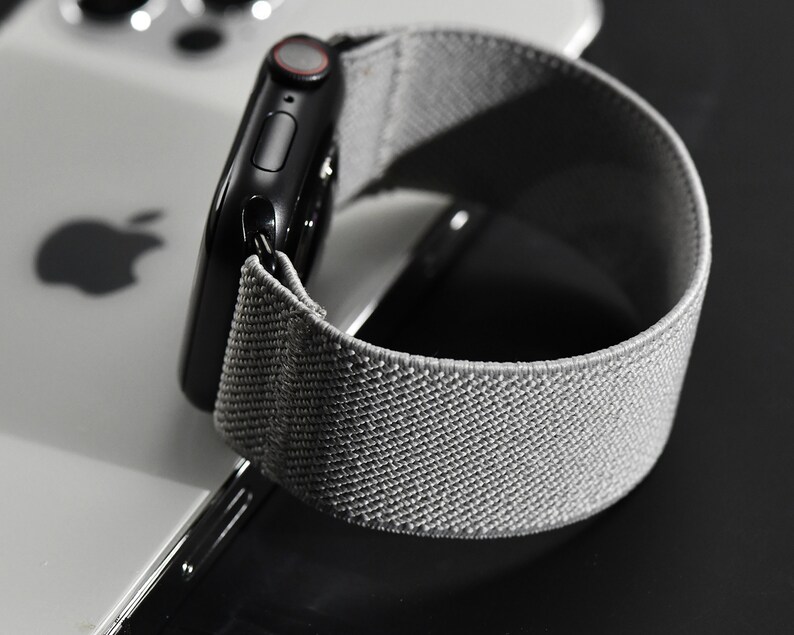 May include: A black Apple Watch with a gray sport loop band. The watch is on a white surface.