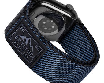 May include: A black Apple Watch with a blue and black woven watch band. The band has a black label with the text "FORTITUDE BY TEFECA EST. 2018".