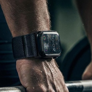 May include: A black smartwatch with a dark grey strap is worn on a wrist. The watch face shows the time as 10:09. The strap appears to be made of a textured, woven material. The watch case is black and rectangular.