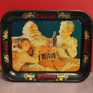 May include: Vintage Coca-Cola serving tray with a black border. The tray features a colorful illustration of Santa Claus with two children, holding a bottle of Coca-Cola. The tray has the Coca-Cola logo and festive decorations.