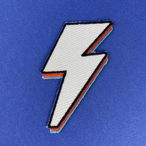 May include: White lightning bolt patch with black, red, and blue trim. The embroidered patch is on a blue background. The bolt is a zig-zag shape, and the patch has a textured surface.
