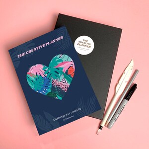 Puede incluir: Un planificador azul con un diseño en forma de corazón con hojas y flores tropicales. El texto "THE CREATIVE PLANNER" está impreso en rosa en la portada. El texto "Challenge your creativity by Doodlemood" está impreso en la parte inferior de la portada.