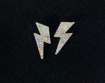 May include: Two silver glitter lightning bolt earrings. The earrings are on a black background.