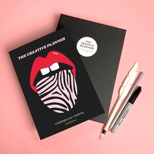 Puede incluir: Un planificador negro con una ilustración de una lengua de cebra roja y blanca. El planificador se titula "The Creative Planner" y tiene el texto "Challenge your creativity by Doodlemood" en la parte inferior.