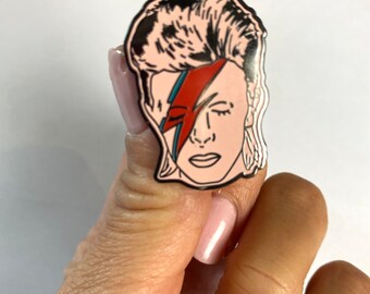 Puede incluir: Un pin de esmalte plateado con un dibujo en blanco y negro de la cara de David Bowie con un rayo rojo y azul sobre su ojo.