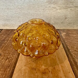 May include: Amber-colored glass bowl with a textured, bubble-like design. The bowl has a rounded shape with a small opening at the top. The glass has a warm, golden hue, and the surface is covered in small, rounded bumps.
