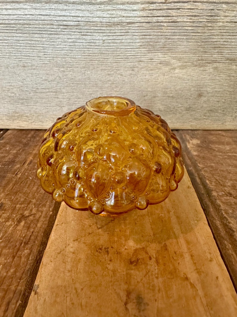 May include: Amber-colored glass bowl with a textured, bubble-like design. The bowl has a rounded shape with a small opening at the top. The glass has a warm, golden hue, and the surface is covered in small, rounded bumps.