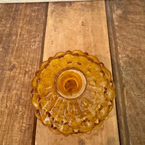 May include: Amber-colored glass bottle stopper with a textured, floral-like design. The stopper has a round shape with a central opening. The glass has a glossy finish and is set against a wooden background.