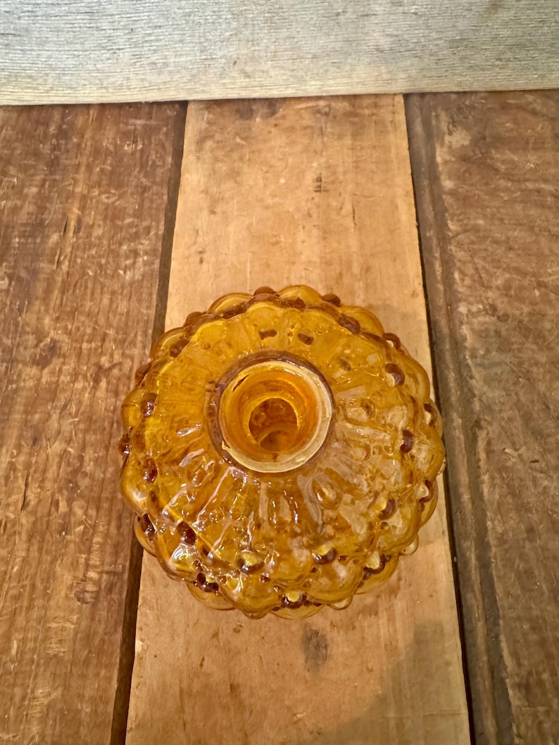 May include: Amber-colored glass bottle stopper with a textured, floral-like design. The stopper has a round shape with a central opening. The glass has a glossy finish and is set against a wooden background.