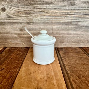 May include: A white ceramic sugar bowl with a matching lid and a small silver metal spoon resting inside. The container features a cylindrical body with a slightly flared rim and a rounded knob handle on top of the lid, presenting a smooth finish.