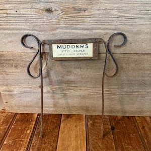May include: A vintage metal foot scraper with the text "Mudders Little Helper Safety Foot Scraper" on a rectangular sign. The scraper has a rusty brown finish and a decorative scroll design on the sides.