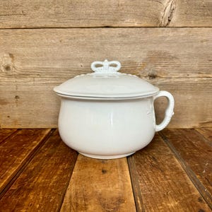 May include: A white ceramic chamber pot with a lid, handle, and crown-shaped finial. The pot has a rounded shape and a decorative floral pattern. The background is a wooden surface.