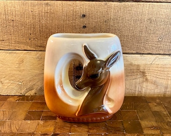 May include: A ceramic vase with a brown and white glaze. The vase features a deer head design in relief. The vase is sitting on a wooden surface.