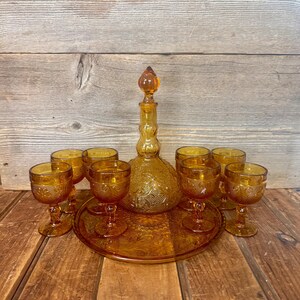 May include: Amber-colored glass drinkware set. The set includes a decanter with a stopper, a serving tray, and eight stemmed glasses. The glassware features an embossed floral pattern. The set is displayed on a wooden surface.