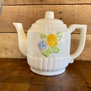 May include: White ceramic teapot with a floral design featuring yellow, blue, and green flowers. The teapot has a spout and a handle.