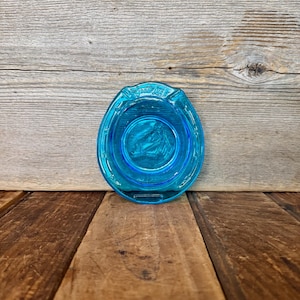 May include: A turquoise glass ashtray in a horseshoe shape. The ashtray has a circular indentation in the center with a horse image. The rim of the ashtray has the text "Lucky Strike".