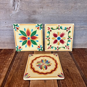 May include: Three square ceramic coasters with floral designs. Each coaster features a cream-colored background with a central flower in red, blue, and green. The coasters are arranged on a wooden surface.