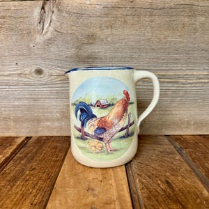 May include: Cream-colored ceramic pitcher with a blue rim and handle. The pitcher features a pastoral scene of a rooster, chicks, and a barn. The rooster has red, brown, and white feathers, and a blue tail. The pitcher is a farmhouse style.