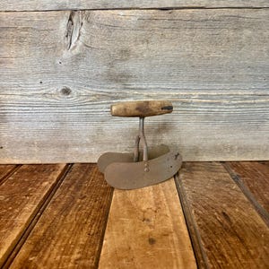 May include: An antique, rusty metal food chopper with a wooden handle. The chopper has a curved blade and a handle that is attached to the blade with a metal rod. The chopper is sitting on a wooden surface.