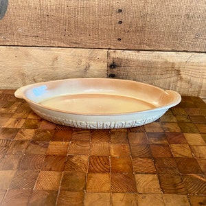 May include: A light beige oval ceramic baking dish with two handles. The dish is sitting on a wooden chopping board.