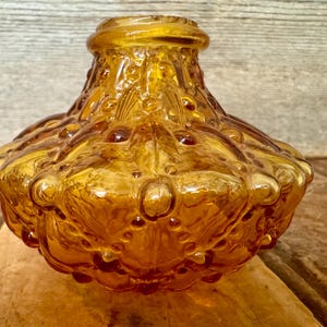 May include: A vintage amber glass bottle with a textured, bubble-like design. The bottle has a wide, rounded base and a narrow neck. The glass has a glossy finish and is set against a wooden background.