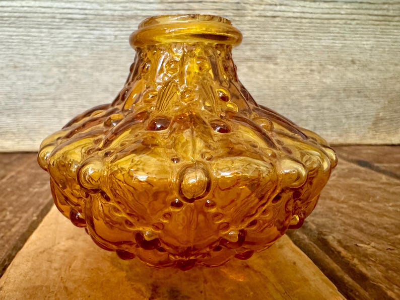 May include: A vintage amber glass bottle with a textured, bubble-like design. The bottle has a wide, rounded base and a narrow neck. The glass has a glossy finish and is set against a wooden background.