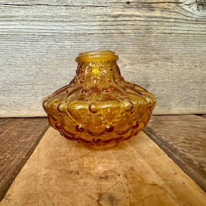 May include: Amber-colored glass vase with a textured, bubble-like pattern. The vase has a rounded body and a slightly flared rim. It sits on a wooden surface against a wooden background. The vase is likely vintage or retro.