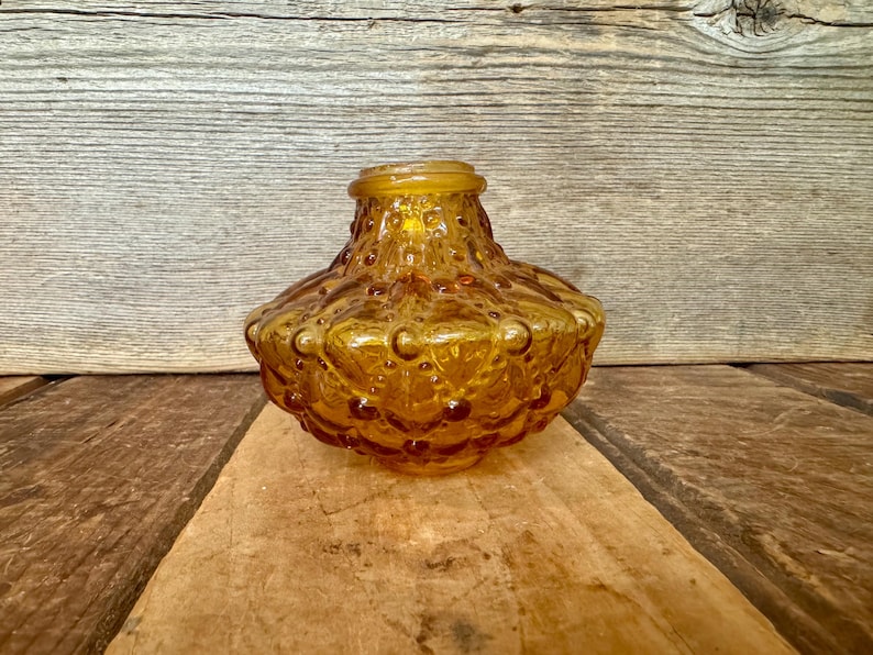 May include: Amber-colored glass vase with a textured, bubble-like pattern. The vase has a rounded body and a slightly flared rim. It sits on a wooden surface against a wooden background. The vase is likely vintage or retro.