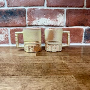 Peut inclure: Deux mugs en céramique beige avec des poignées carrées et des détails décoratifs. Les mugs ont une forme cylindrique avec une surface texturée et sont posés sur un fond de mur en briques. Les mugs sont sur une surface en bois.