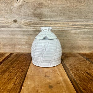 May include: A white ceramic honey pot shaped like a traditional skep beehive. The container features a textured, horizontal ribbed pattern resembling woven straw and includes a matching lid with a small notch for a spoon to rest inside.
