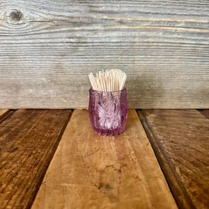 May include: A small, ornate, pink glass toothpick holder filled with wooden toothpicks. The holder has a textured, floral design and sits on a wooden surface. The toothpicks are visible at the top of the holder.