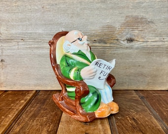 May include: A ceramic figurine of an elderly person in a rocking chair, reading a newspaper. The figure wears a green robe, glasses, and has a white beard. The newspaper reads "RETIR FU". The chair is brown with a yellow back.