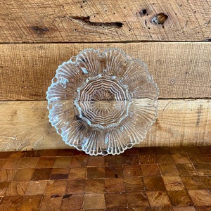 May include: A clear glass plate with a scalloped edge and a decorative centre design. The plate is on a wooden surface.