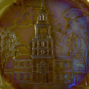 Peut inclure: Une assiette en verre iris&eacute; vintage avec une image en relief de l'Independence Hall &agrave; Philadelphie, en Pennsylvanie. L'assiette est de couleur dor&eacute;e avec une l&eacute;g&egrave;re teinte violette.