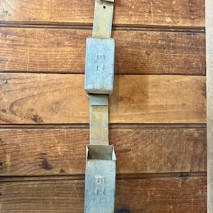May include: A vintage metal wall-mounted mail holder with a weathered, aged appearance. The rectangular compartments are connected by a strip of metal. The letters "EZ" are visible on the front of the compartments. The mail holder is mounted on a wooden wall.
