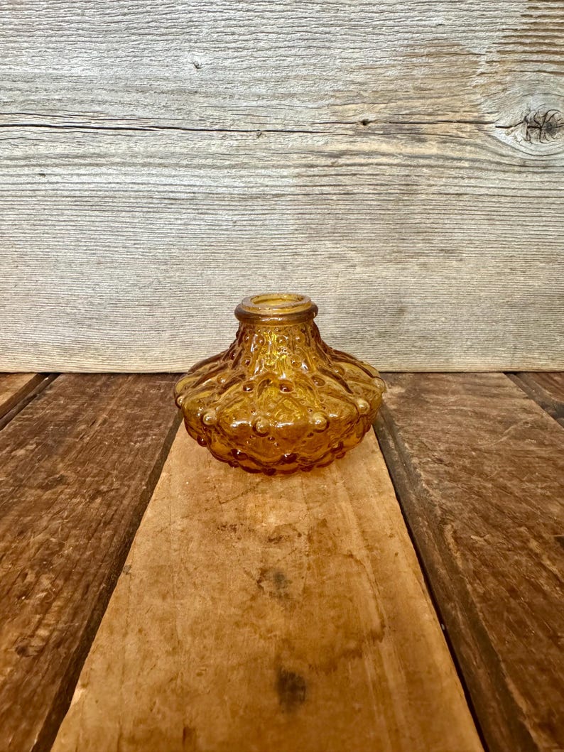 May include: Amber-colored glass decorative vase with a textured, bubble-like design. The vase has a round base and a flared top. The glass has a warm, golden hue, and the surface is covered in small, raised bumps.