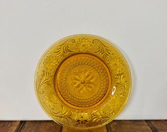 May include: A round, amber-colored glass plate with a floral design. The plate features a central flower motif surrounded by intricate floral and scrollwork patterns. The plate is made of transparent glass.