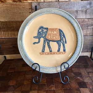 May include: A ceramic plate with a blue and brown elephant design. The elephant is wearing a saddle with the words "HAN I BLE" written on it. The plate has a light blue rim and a beige background.