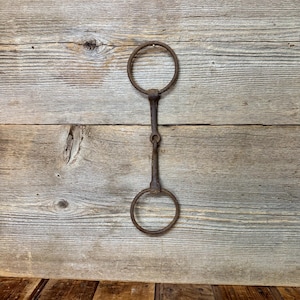 May include: A weathered, dark brown metal horse bit featuring two circular rings connected by a central jointed bar. The object displays a heavily oxidized, rustic texture throughout its entire surface, showing signs of age and consistent wear.