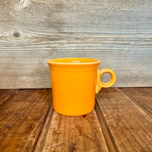 May include: A bright yellow ceramic mug with a rounded handle. The mug is sitting on a wooden surface.