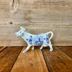 May include: A white ceramic cow figurine with blue floral and windmill designs. The cow has a curved handle and is posed in a walking position. The figurine is displayed on a wooden surface.