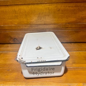 May include: A vintage white Frigidaire Hydrator with a rectangular shape and a lid. The metal container has black trim and some signs of wear, including rust spots. The text "Frigidaire Hydrator" is printed on the front.