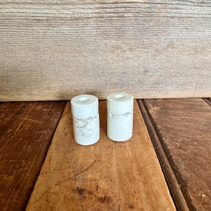May include: Two white marble candle holders with a rough, textured surface. The candle holders are cylindrical in shape and are sitting on a wooden surface.