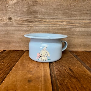 May include: A light blue enamel chamber pot featuring a painted illustration of a small cream-colored bunny with a pink butterfly on its side. The vessel has a wide, flat rim and a single side handle, with a smooth, glossy finish throughout.